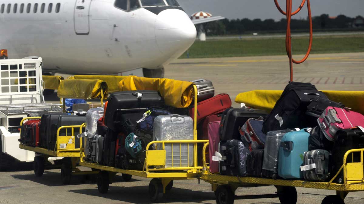 Bilde av en rekke kofferter som står stablet på transportbil på vei mot et fly på flyplassen. 